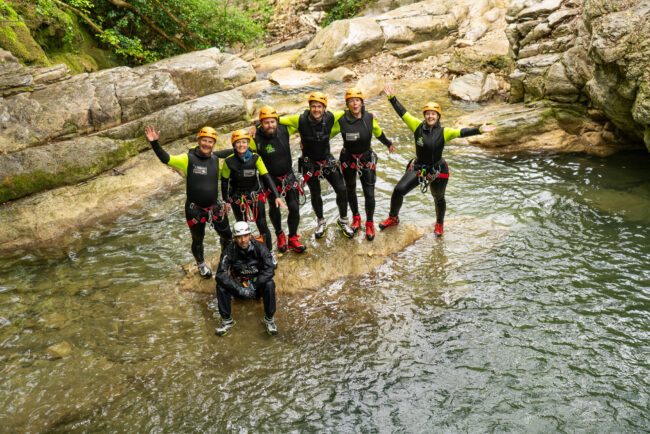 Gruppo di partecipanti durante un’esperienza di canyoning al Rio Buti a Vaiano, in provincia di Prato, Toscana. Sei persone con casco e muta tecnica da canyoning posano sorridenti su una roccia al centro del torrente, circondate da pareti rocciose levigate dall’acqua e vegetazione verde tipica dell’Appennino toscano. Attività outdoor in Val di Bisenzio, vicino Prato, ideale per chi cerca canyoning in Toscana.
