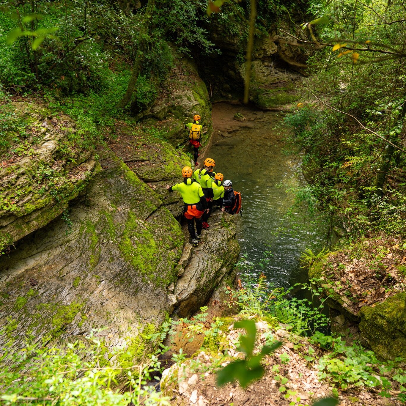 Gruppo di canyoning che avanza lungo una stretta gola del Rio Buti a Vaiano, Prato, nel cuore della Toscana. I partecipanti, equipaggiati con casco, imbracatura e muta in neoprene, camminano tra rocce stratificate e acqua corrente bassa, immersi in un canyon verde e selvaggio della Val di Bisenzio. Percorso di canyoning in Toscana ideale per avventura e natura.